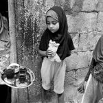 three people appear in a black and white photo. from left to right is a woman in a hijab offering tea to two girls. one girl in the middle eats bread and looks at the tea. the girl on the far right looks off into the distance.