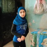 a girl in a blue Hijab stands in front of a domestic space. she wears a blue shirt with a silver bow. in the top right corner is gold and pink fabric in the shape of a bow.