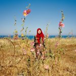 a girl in a red hijab stands in a field of pink flowers. her eyes are closed. behind her is the open ocean