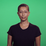 A woman with glasses and a black t-shirt stands in front of a green screen, pictured from the ribcage up.
