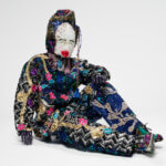 Sculpture of a seated person encrusted with colorful beads, bells, sequins, and other homespun materials.
