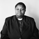 A black-and-white photograph of a person from the waist up wearing a blazer and clergy shirt against a blank wall