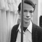 A black and white photo shows a man in a bathroom. The image is vertically split by what appears to be a gap between two panels of a mirrored medicine cabinet door. The man's mouth is curled in a half smile but he does not look happy. His face is thin and he wears a thick cardigan.