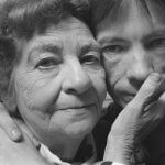 A black and white photo shows a woman with a worn face pressed close with that of her son's, which is gaunt. Her hand touches his cheek. His hands are clasped together on her shoulder.
