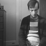 A black and white photo shows a thin man with protruding cheekbones sitting with his face turned down. There's a wooden dresser to the left of the image and he's wearing a thick cardigan over another sweater and jeans. There's a closed door behind him.