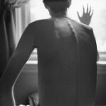A black and white photo shows the shirtless back of a very thin man sitting in front of a bright window. His right hand is on the glass as he looks out. His spine and shoulder blades protrude.